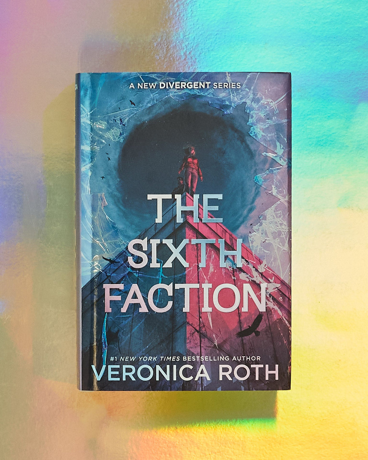 The book cover of 'The Sixth Faction' by Veronica Roth, displayed upright against a colorful iridescent background. The cover features a dark blue circular portal with a figure standing on a red platform, and the title is written in large white letters.